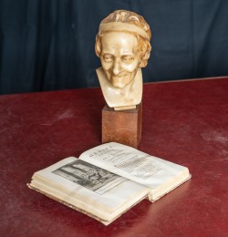 die wachsähnliche Büste Voltaires auf einem Sockel; er scheint, auf sein eigenes Buch hinabzublicken, das aufgeschlagen vor ihm liegt                                                         