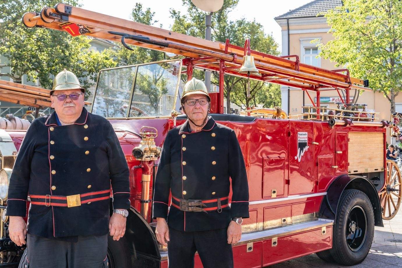 zwei Mitglieder der Alterswehr in historischer Uniform und mit goldglänzendem Helm vor ihrem roten Feuerwehr-Oldtimer                                    